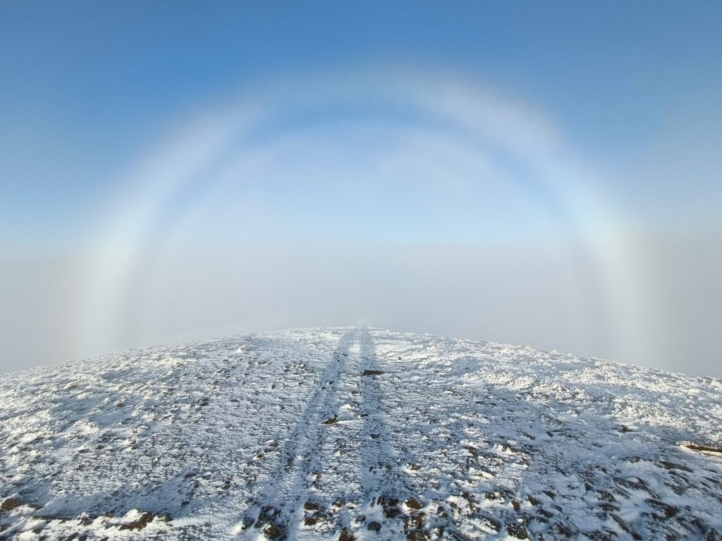 Snow covered ground with the long shadows of two people standing off camera. A white fog-bow of cloud on blue sky frames the shadows.